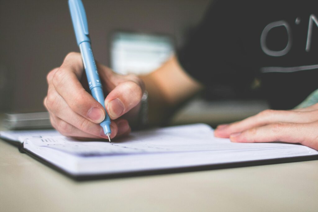 Close-up of hand writing in notebook using a blue pen, focus on creativity. Best IELTS Coaching Online